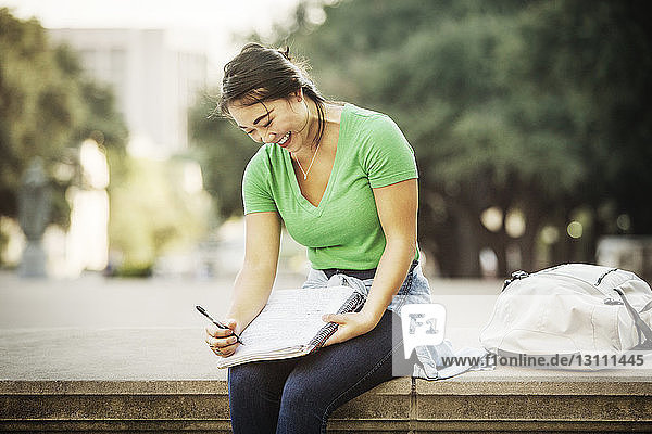 Glückliche Frau schreibt in Buch  während sie auf Stützmauer sitzt
