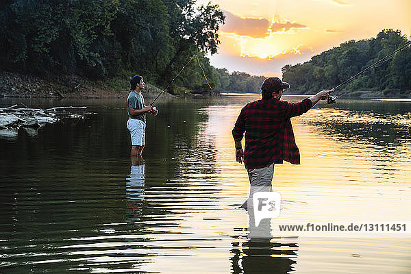 Male friends fishing while standing in lake during sunset