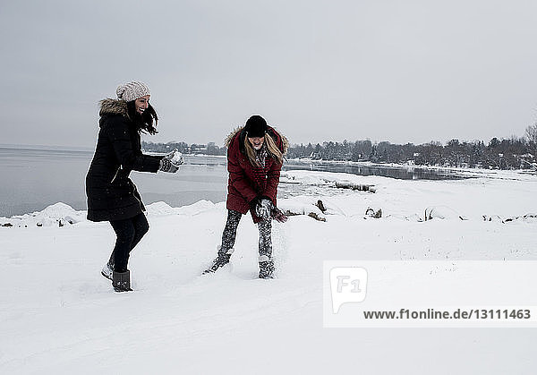 Female friends playing with snow at beach against sky during winter