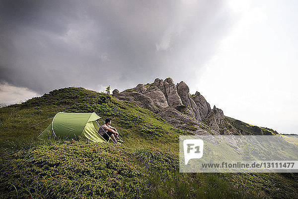 Low angle view of hiker sitting by tent on mountain against cloudy sky