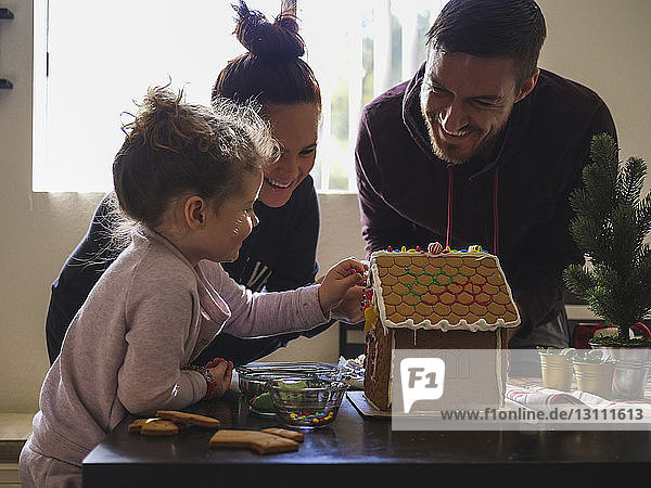 Glückliche Eltern sehen Tochter beim Dekorieren des Lebkuchenhauses auf dem Tisch