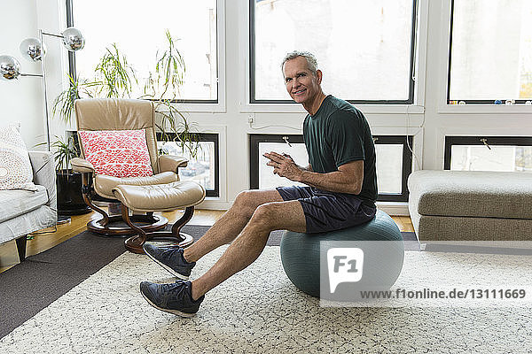 Full length portrait of mature man sitting on fitness ball against windows at home