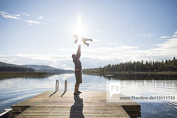 Full length of playful father throwing son in air against lake and sky during sunny day