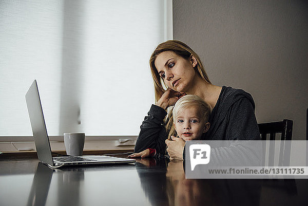 Porträt eines kleinen Jungen  der mit seiner Mutter mit einem Laptop auf einem Tisch zu Hause sitzt