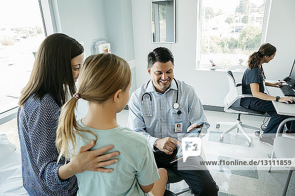 High angle view of pediatrician showing x-ray image on tablet computer to mother and daughter while female doctor working in background