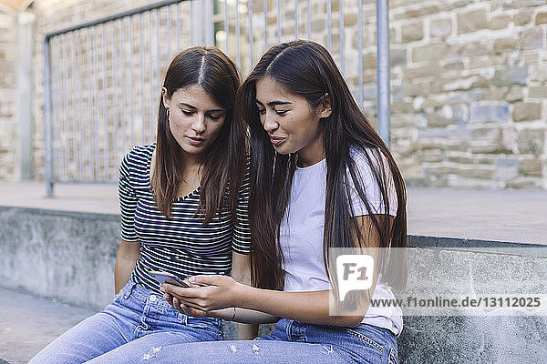 Freunde benutzen Mobiltelefon  während sie auf Stufen an der Wand sitzen