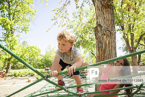 Smiling mother looking at son climbing on ropes in playground