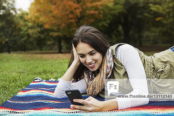 Happy woman using mobile phone while lying on blanket at park