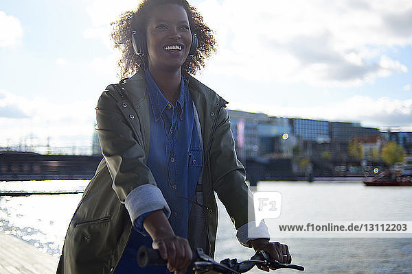 Niedrigwinkelansicht einer glücklichen Frau  die Fahrrad fährt  während sie Musik gegen den Himmel hört