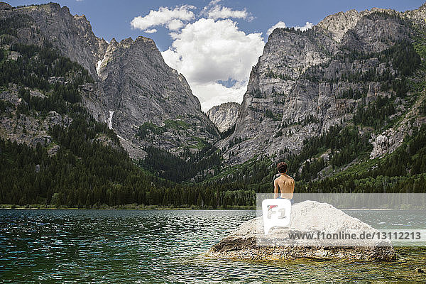 Rear view of teenager boy sitting on rock in lake
