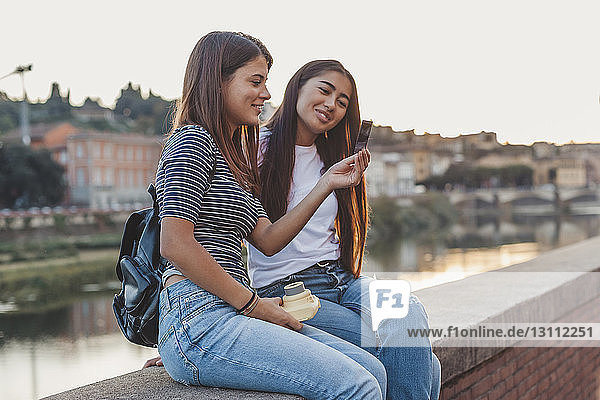 Lächelnde Freundinnen schauen auf die sofortige Druckübertragung  während sie auf einer Stützmauer am Kanal in der Stadt sitzen