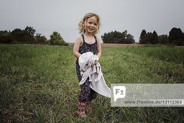Porträt eines glücklichen Mädchens mit Stoff in der Hand  das auf einem Grasfeld vor dem Himmel im Park steht