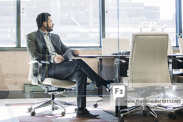 Businessman looking away while sitting in board room seen through glass