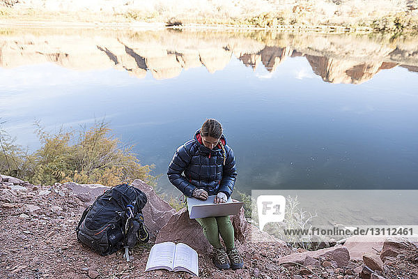 Hochwinkelaufnahme einer Wanderin mit Laptop-Computer  die auf einem Felsen am See sitzt