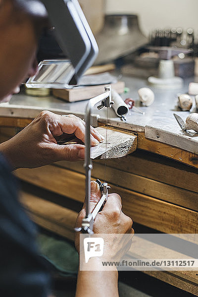 Beschnittenes Bild einer Handwerkerin  die mit einer Handsäge Schmuck auf einem Holztisch in einer Werkstatt herstellt
