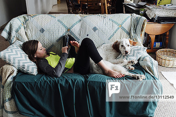 Mädchen benutzt Tablet-Computer  während sie mit Hund zu Hause auf dem Sofa liegt