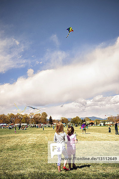 Rear view of siblings flying kites at park against cloudy sky