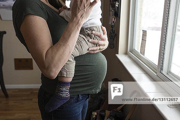 Mittelsektion einer schwangeren Mutter  die ihren Sohn gegen das Fenster trägt