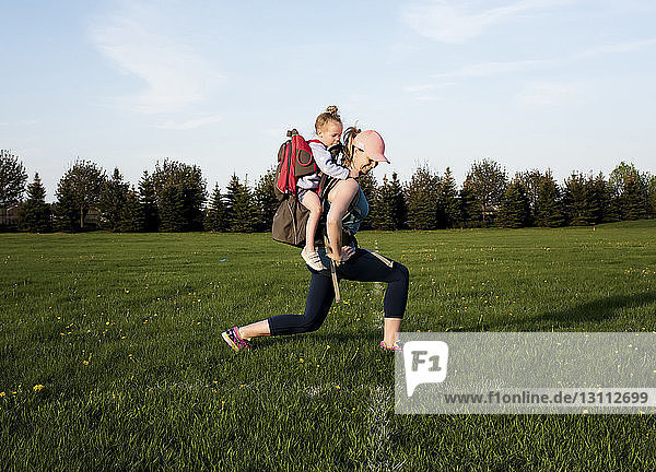 Side view of mother carrying daughter while exercising on grassy field against sky at park