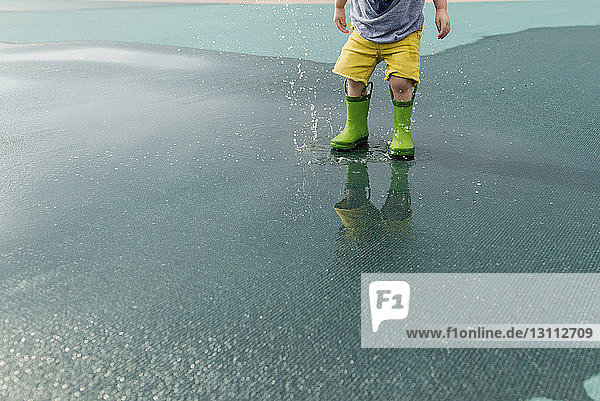 Niederer Teil eines Jungen in Gummistiefeln  der auf nasser Straße Wasser spritzt