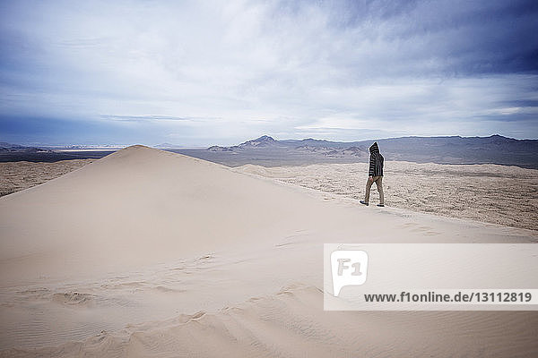 Man walking on sand dune in desert against cloudy sky