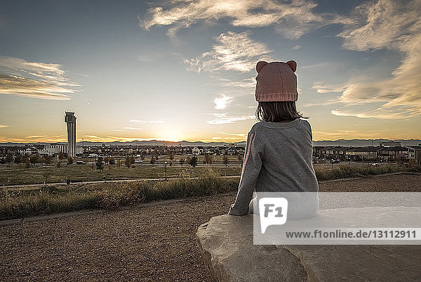 Mädchen betrachtet Stadtlandschaft  während sie bei Sonnenuntergang auf einem Felsen gegen den Himmel sitzt