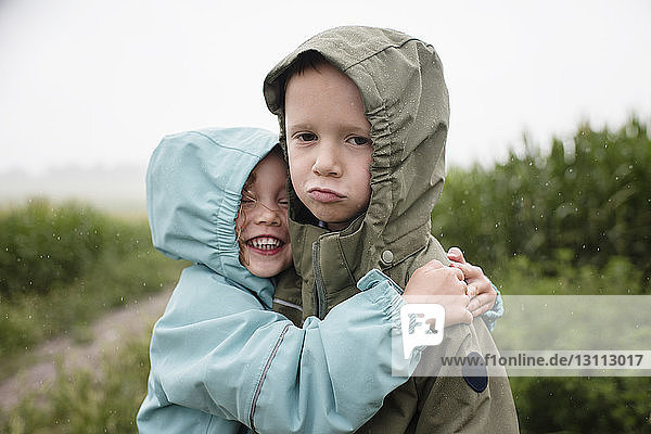 Porträt eines traurigen Bruders  der von einer glücklichen Schwester umarmt wird  während er in der Regenzeit vor Pflanzen steht