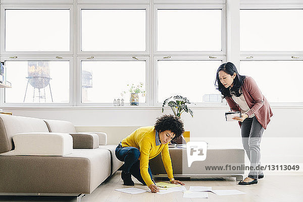 Businesswoman looking at female colleague writing on papers while kneeling in office