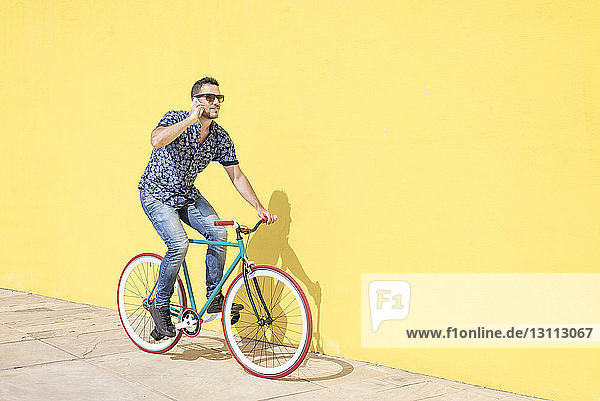 Mann spricht in voller Länge mit einem Smartphone  während er am sonnigen Tag mit dem Fahrrad gegen eine gelbe Wand auf dem Bürgersteig in der Stadt fährt