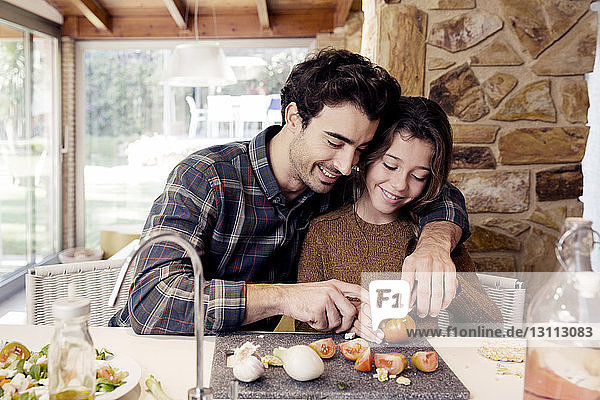 Lächelnder Vater hilft Tochter beim Tomatenschneiden  während er am Tisch sitzt