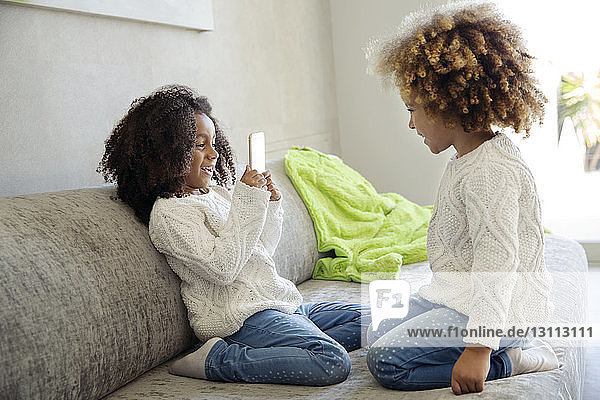 Fröhliches Mädchen fotografiert Schwester mit Smartphone auf Sofa