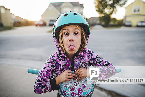 Porträt eines verspielten Mädchens mit herausgestreckter Zunge beim Rollerfahren auf dem Fussweg