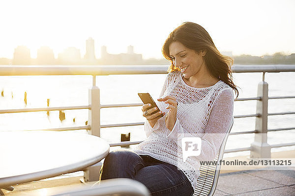 Glückliche Frau benutzt Smartphone im Café an der Promenade während des Sonnenuntergangs