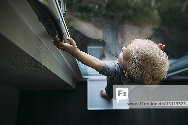Hochwinkelansicht eines kleinen Jungen  der durch ein Fenster schaut  während er zu Hause auf einem Hocker steht