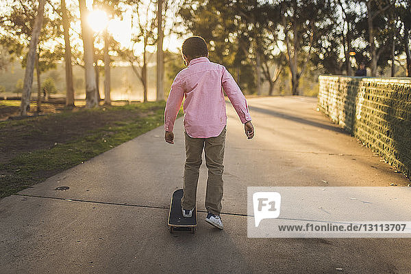 Rückansicht eines Jungen  der bei Sonnenuntergang auf einem Fußweg im Park Skateboard fährt