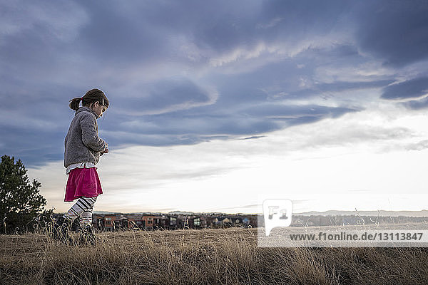 Niedrigwinkelaufnahme eines Mädchens in warmer Kleidung  das bei Sonnenuntergang auf einem Grasfeld vor bewölktem Himmel geht