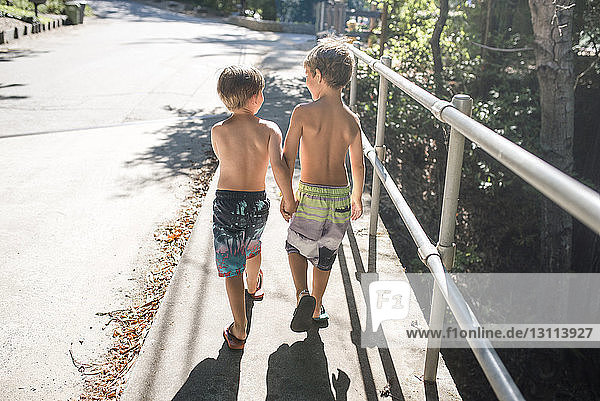 Rear view of shirtless brothers holding hands while walking on footpath by railing during summer