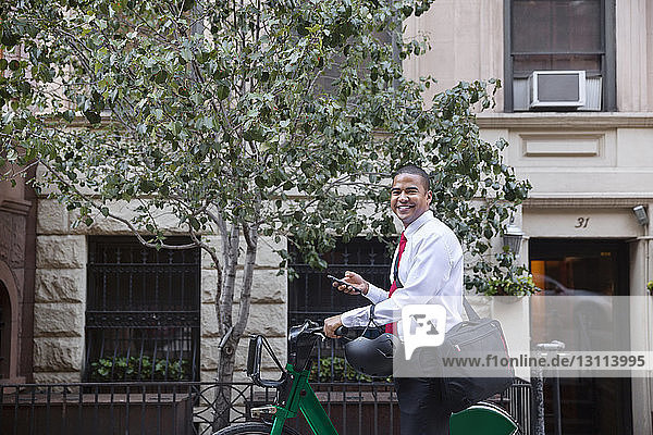 Porträt eines lächelnden Geschäftsmannes mit Fahrrad  der ein Mobiltelefon benutzt  während er gegen ein Gebäude steht