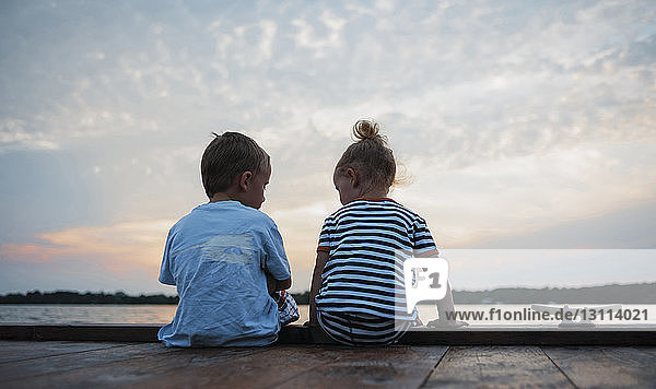 Rear view of siblings sitting on pier against cloudy sky during sunset