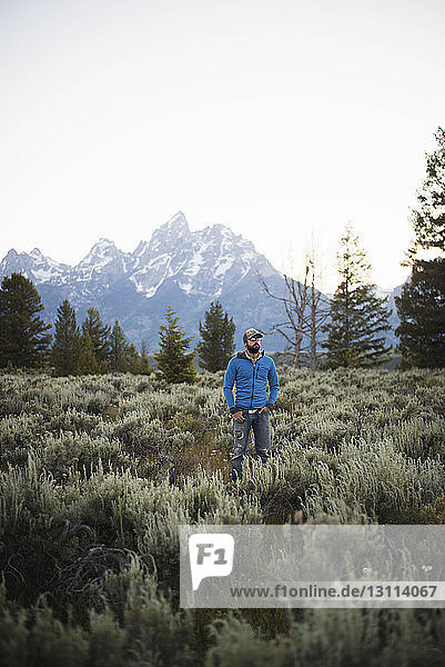 Hiker with hands in pockets standing on field against mountains and clear sky at Grand Teton National Park