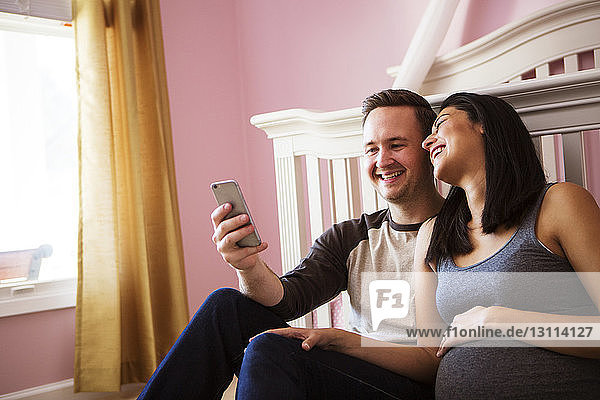 Glücklicher Mann benutzt Smartphone  während er zu Hause neben schwangerer Frau sitzt