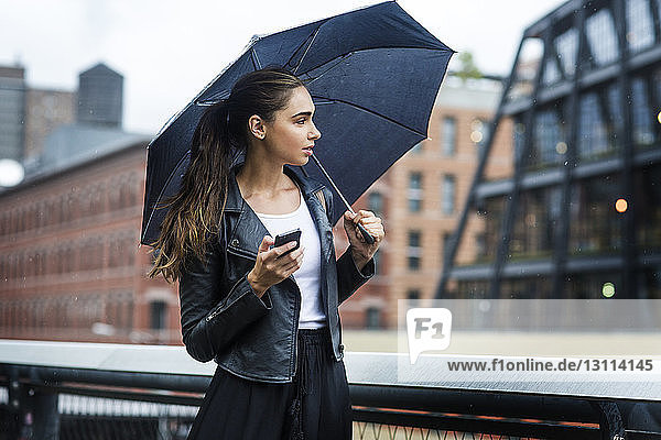 Woman carrying umbrella and using phone while standing at railing in city