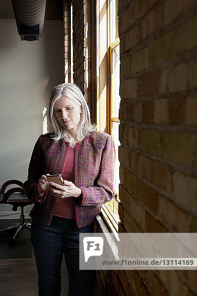 Reife Geschäftsfrau nutzt Smartphone am Fenster im Kreativbüro