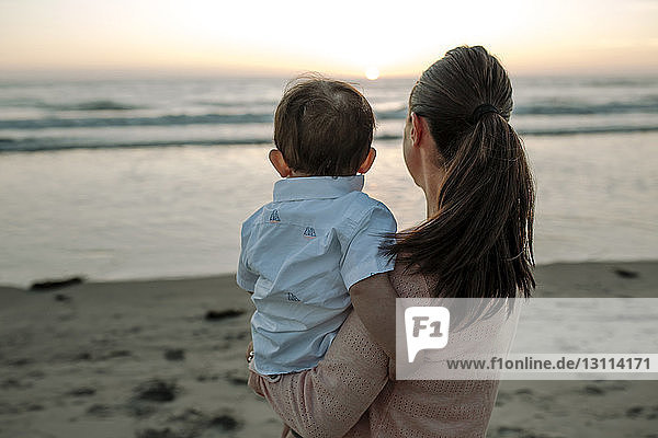 Seitenansicht einer Mutter  die einen Sohn trägt  während sie bei Sonnenuntergang am Strand gegen den Himmel steht