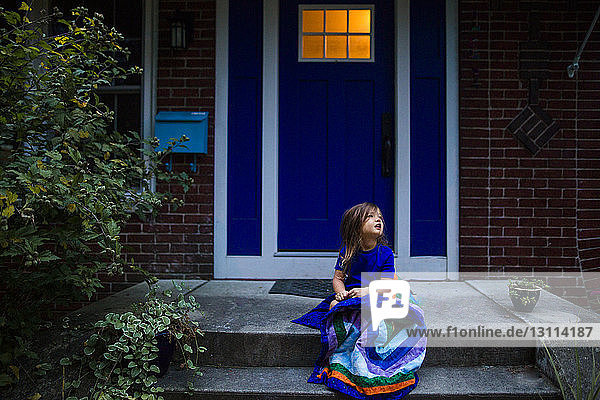 Girl with blanket sitting on porch while looking away against house