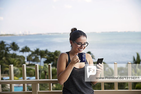 Happy woman using smart phone while drinking coffee against sea