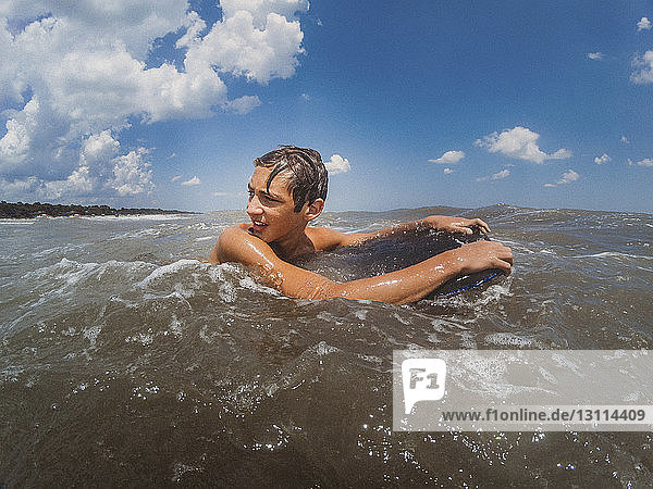 Shirtless teenage boy looking away while surfing on sea against blue sky