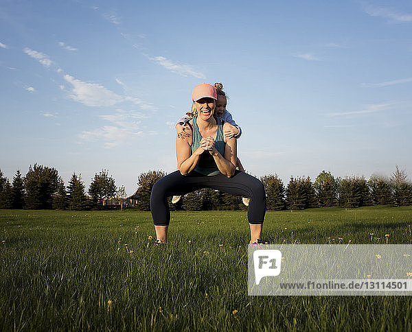 Glückliche Mutter trägt ihre Tochter  während sie auf einem Grasfeld im Park gegen den Himmel trainiert