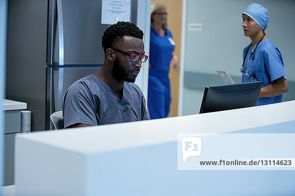 Confident doctor using desktop computer at hospital reception with colleagues working in background