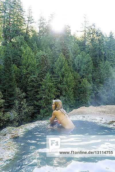 Frau im Bikini schaut weg  während sie sich in einer heißen Quelle im Wald entspannt